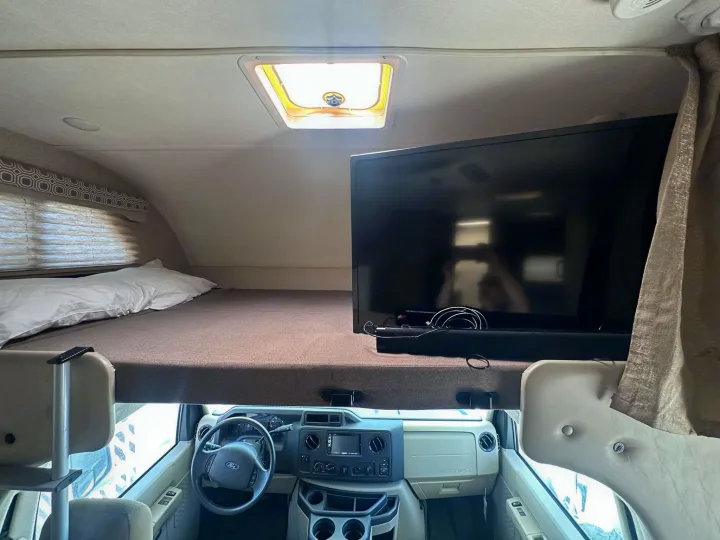 Cozy RV rental interior: overhead bunk bed with TV, showing the driver's cab and dashboard below.. Thor Motor Coach Four Winds 2019