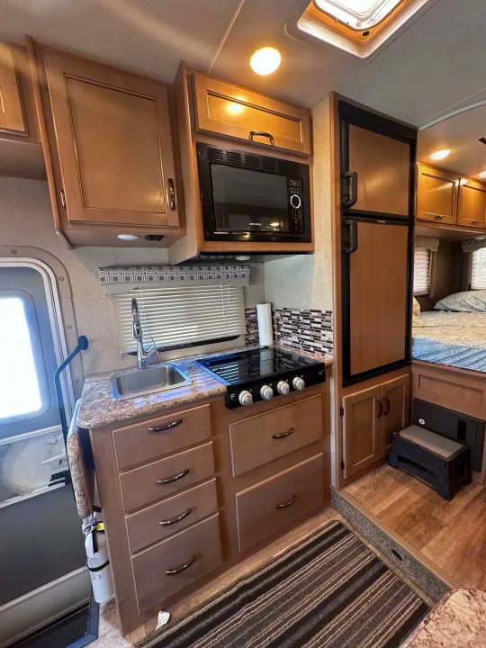 Cozy RV rental interior featuring a full kitchen with microwave, stove, sink, and fridge, plus a glimpse of the bed.. Thor Motor Coach Four Winds 2019