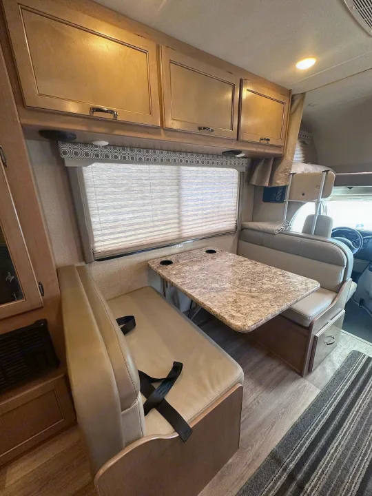 View of an RV rental's dinette area featuring a table, upholstered benches with seatbelts, overhead cabinets, and a window.. Thor Motor Coach Four Winds 2019