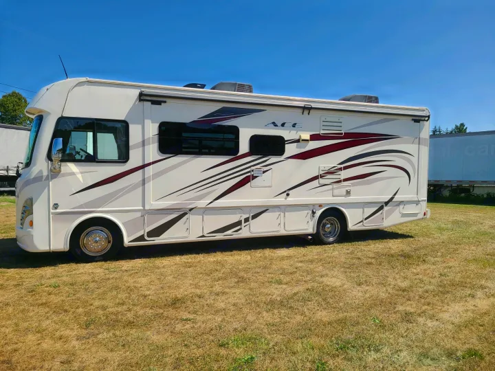 Explore the open road in this clean white RV rental, featuring sleek graphics. Parked on a grassy field under a clear blue sky.. Thor Motor Coach A.C.E 2018