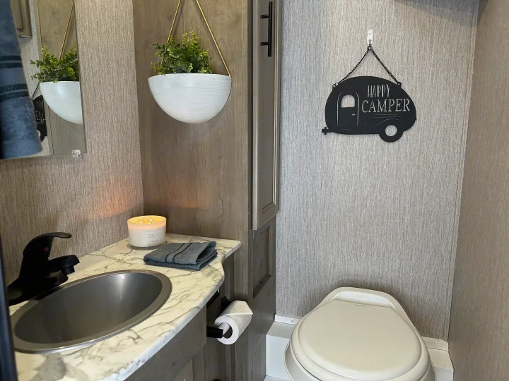 This clean RV rental bathroom features a modern sink, toilet, mirror, and decor like a hanging plant and "Happy Camper" sign.. Forest River Coachmen Freelander 2022