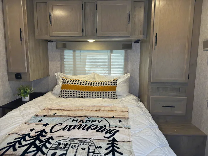 Cozy RV rental bedroom featuring a clean bed, "Happy Camping" blanket, and light wood-look storage cabinets.. Forest River Coachmen Freelander 2022