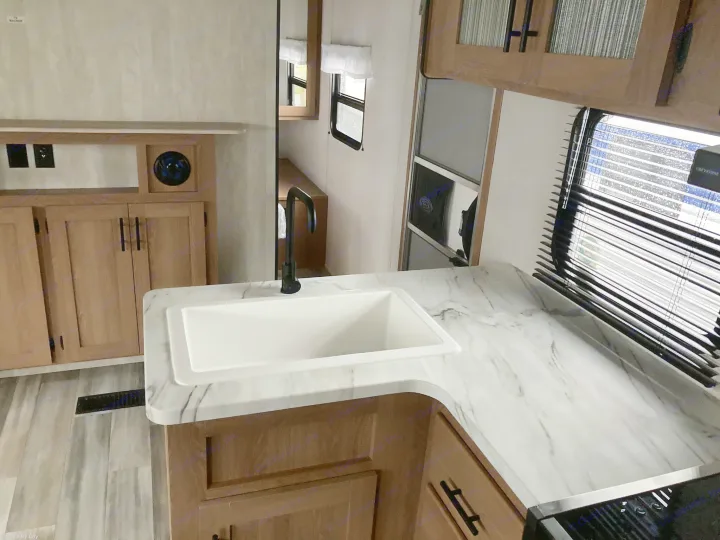 View of a clean RV rental kitchen with a white sink, black faucet, and marble-patterned countertops over light wood cabinets.. Forest River Avenger LE 2022