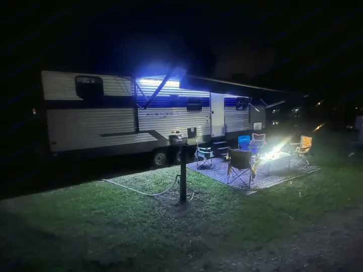 Nighttime rv rental camping setup: a travel trailer with bright blue awning lights and an outdoor seating area.. Forest River Avenger LE 2022