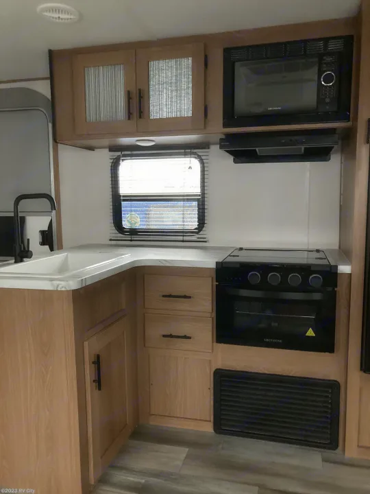 Detailed view of an RV rental kitchen: L-shaped counter, sink, microwave, stove/oven, wood cabinets, and window.. Forest River Avenger LE 2022