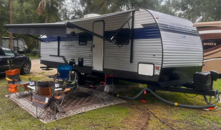 This RV rental travel trailer is set up at a green campsite with an extended awning, outdoor rug, chairs, and table.. Forest River Avenger LE 2022