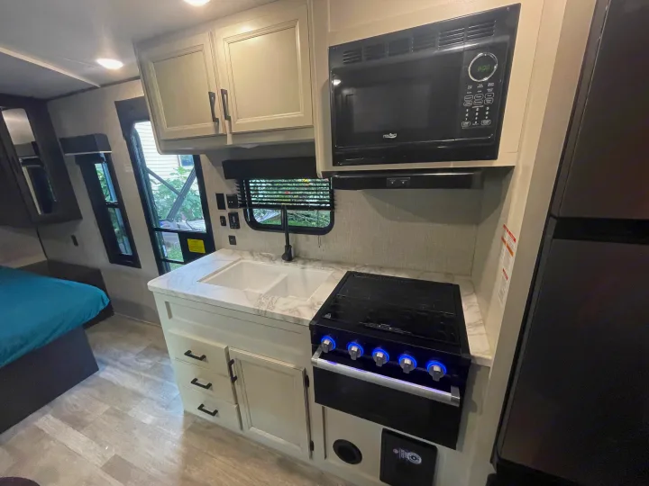 RV rental interior: A modern kitchen area featuring a microwave, stove, sink, and cabinetry, with a bed visible.. Jayco Jay Flight 2023