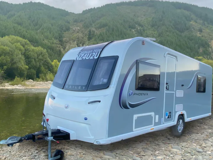 A modern RV rental (caravan) is parked by a scenic river, nestled against lush green mountains, offering tranquil travel.. Bailey Pheonix 640 2022