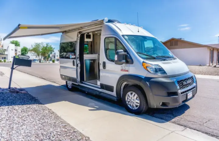 Silver RV rental parked with its side door open, revealing the interior, and awning extended on a sunny residential street.. Thor Rize 2023