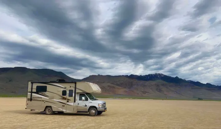 A tan RV rental rests on a vast, flat dry lake bed. Behind it are distant mountains and a dramatic cloudy sky.. Winnebago Minnie Winnie 2020