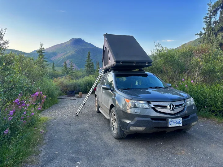 An SUV with a ladder to its open rooftop tent (like an RV rental) at a mountain campsite with a fire pit.. Other Other 2007