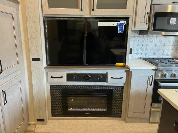 Light-toned RV rental interior with a large smart TV above an electric fireplace, next to a modern kitchen area.. Keystone RV Montana High Country 2023