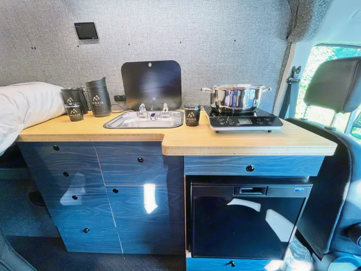 Moxie Van Co. RV rental: a compact camper van kitchen with a sink, induction cooktop, and under-counter refrigerator.. Ford Custom Transit 2022