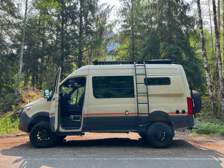 Cream 'MODE 4x4' off-road camper van, an adventure rv rental, parked in a lush forest. Driver door open.. Mercedes-Benz Sprinter 2021