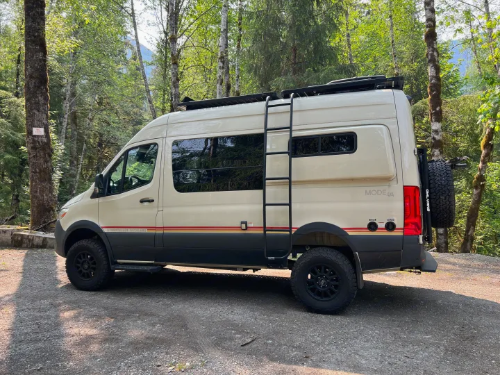 Cream-colored camper van equipped for overland adventures, parked on gravel in a sunny forest. Ideal for rv rental trips.. Mercedes-Benz Sprinter 2021