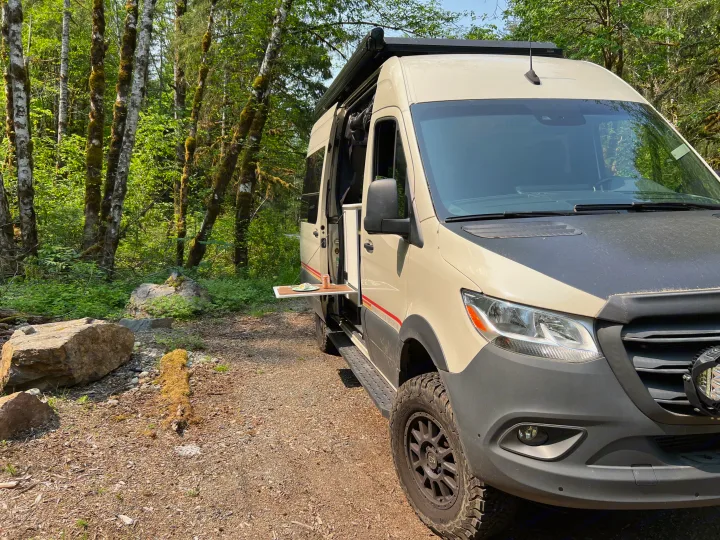 Explore nature with an RV rental! This camper van is parked in a lush forest with a side table extended for a meal.. Mercedes-Benz Sprinter 2021