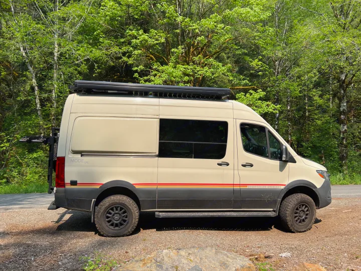 A cream-colored Storyteller Overland 4x4 camper van, ideal for RV rental adventures, parked by a lush forest.. Mercedes-Benz Sprinter 2021