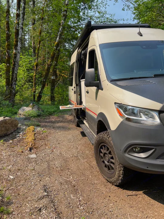 RV rental camper van parked in a forest. Side door open, revealing a pull-out table with a drink and plate.. Mercedes-Benz Sprinter 2021
