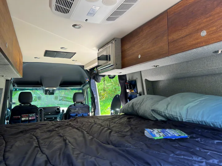 Inside a well-equipped RV rental: a comfy bed, overhead storage, microwave, and front cab view.. Mercedes-Benz Sprinter 2021