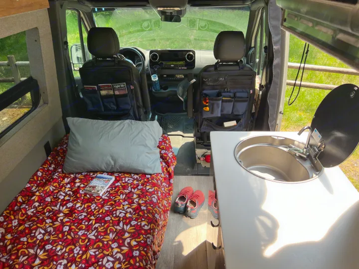 Inside an RV rental: a cozy bed with colorful bedding, a functional kitchen sink, and organized front seats, ready for adventure.. Mercedes-Benz Sprinter 2021