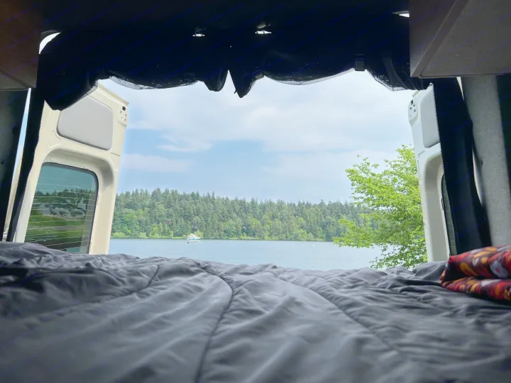 Serene RV rental view from inside: Open van doors showcase a tranquil lake, lush forest, and boat from the comfort of bed.. Mercedes-Benz Sprinter 2021