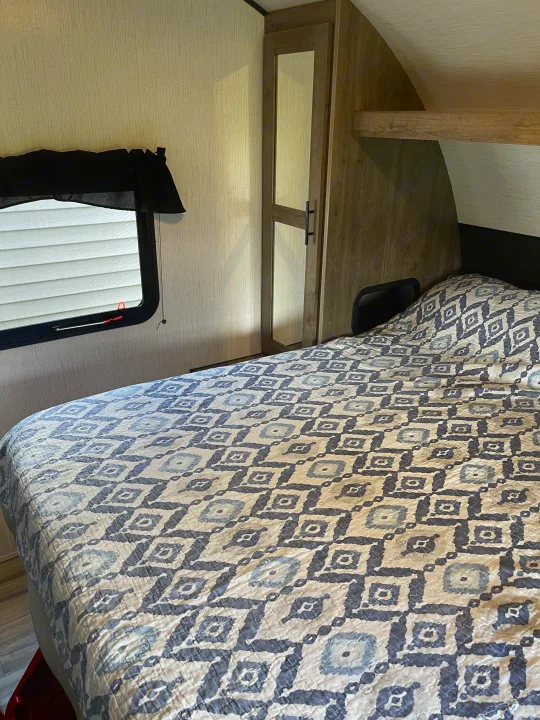 An RV rental bedroom, showcasing a patterned bed, window with blinds, and a built-in mirrored cabinet.. Heartland RVs Pioneer 2022