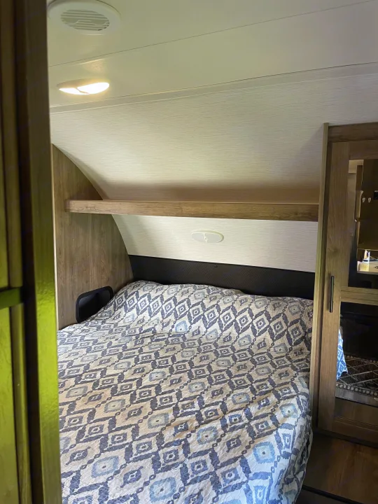 Inside an RV rental: a cozy bedroom featuring a patterned bed, overhead shelf, and wardrobe. Compact, functional design.. Heartland RVs Pioneer 2022