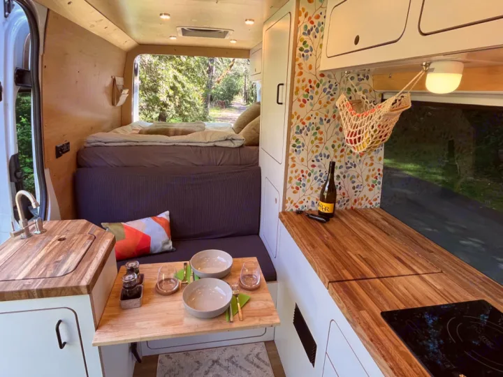 Step inside this inviting RV rental: a camper van interior with a kitchen, dining table, and bed, set against a natural backdrop.. Mercedes-Benz Sprinter 2021