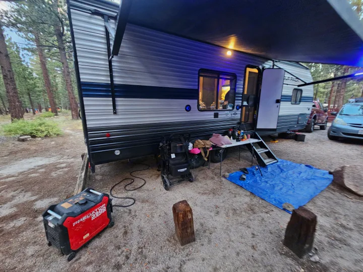 An RV rental is set up for camping in the woods, featuring an extended awning, outdoor table, generator, and supplies.. Forest River Cherokee Wolf Pack 2021