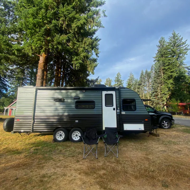 An RV rental: a dark travel trailer is set up on a grassy campsite with two chairs among tall trees.. Fleetwood Prowler 1996