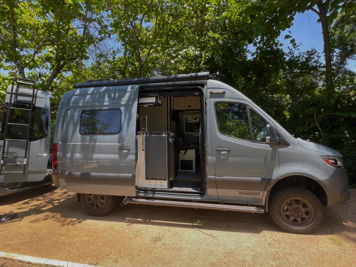 Explore in a Winnebago camper van RV rental. Side door open, revealing a functional interior amidst green trees.. Winnebago Revel 2023