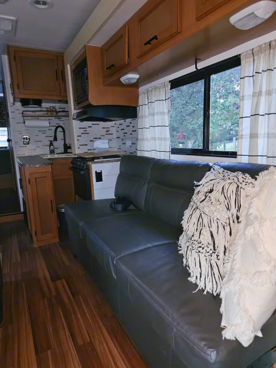 RV rental interior: a compact kitchen with wood cabinets, sink, and microwave, next to a dark leather sofa with cozy throws.. Forest River Coachmen Freelander 2014