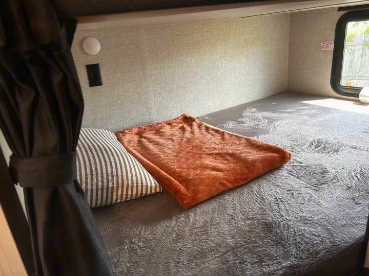 Cozy RV rental bunk bed with soft grey blankets, striped pillow, and an orange throw, ideal for comfortable travel.. Jayco Jay Flight 2023