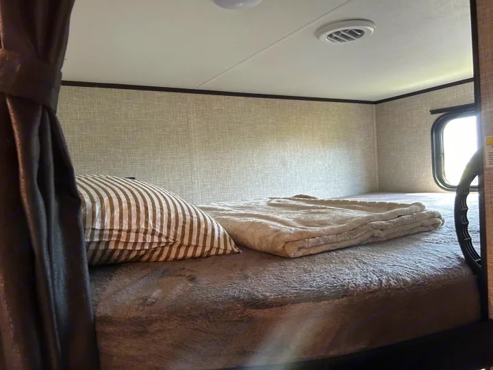 Cozy sleeping area in an RV rental, with a striped pillow, plush blanket, and natural light from the window.. Jayco Jay Flight 2023
