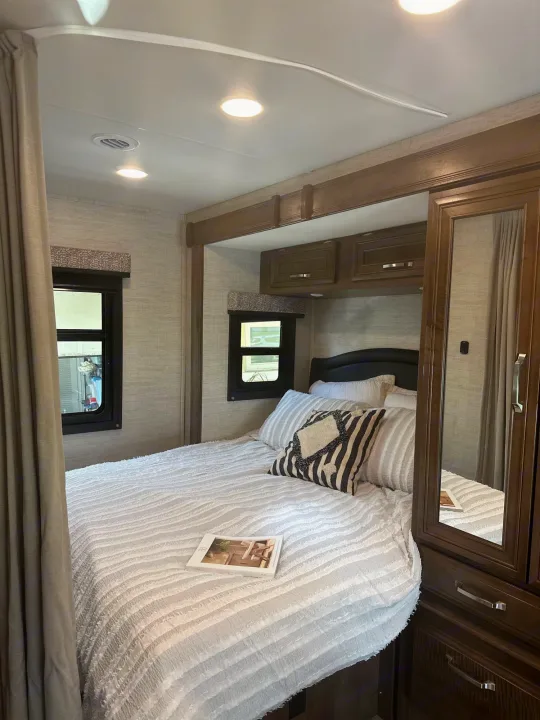 Inside an RV rental: a comfy bedroom with striped and white bedding, dark wood cabinets, windows, and ceiling lights.. Jayco Melbourne 2022