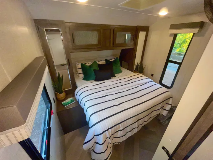Cozy RV rental bedroom featuring striped bedding, overhead cabinets, and a window with a green outdoor view.. Forest River Wildwood 2021