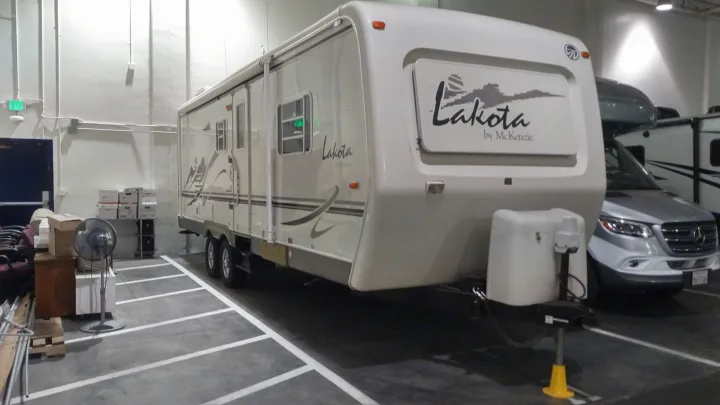 A large white travel trailer, branded 'Lakota by McKenzie', is showcased as an RV rental in a spacious indoor storage.. Other Other 2002