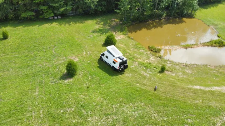 A white RV rental camper van is parked in a vast green field next to a pond with ducks, surrounded by trees.. Winnebago Solis 2023
