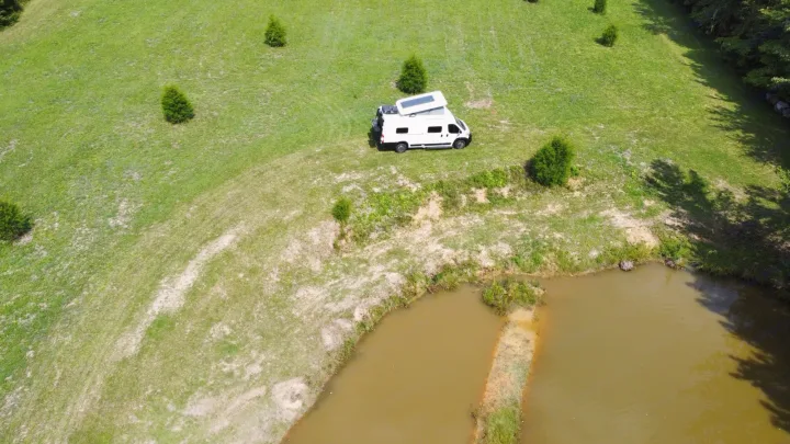 Experience nature with an RV rental! A white camper van with solar panels rests on a green hill beside a tranquil pond.. Winnebago Solis 2023