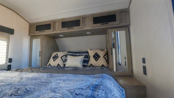 Cozy RV rental bedroom featuring a patterned bedspread, decorative pillows, and compact overhead storage with mirrored cabinets.. Forest River Cherokee Alpha Wolf 2021