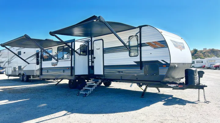 White and black Wildwood travel trailer RV rental with extended awnings & steps, parked on gravel under a clear sky.. Forest River Wildwood 2022