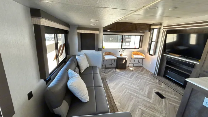 Spacious RV rental living room features a comfy grey sofa, bar stools, TV, fireplace, and modern herringbone flooring.. Forest River Wildwood 2022
