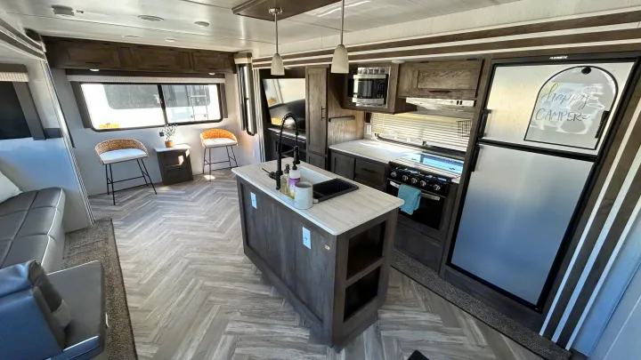 Spacious rv rental interior features a modern kitchen island, stove, microwave, fridge, and living area with herringbone floors.. Forest River Wildwood 2022