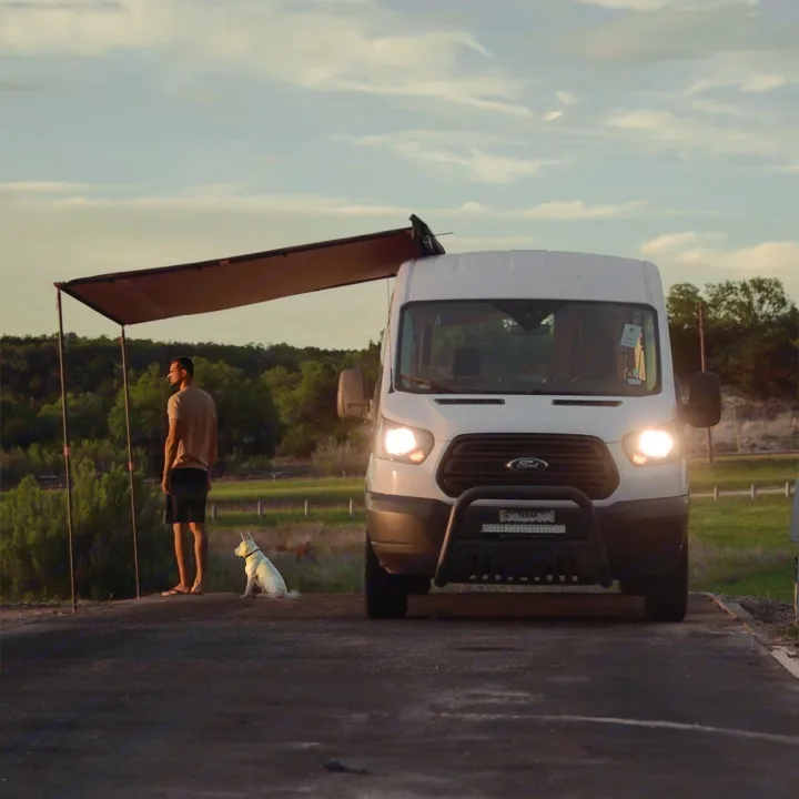 RV rental journey: A man and his white dog relax under the extended awning of a camper van at dusk, enjoying the peaceful outdoors.. Ford Transit 2019