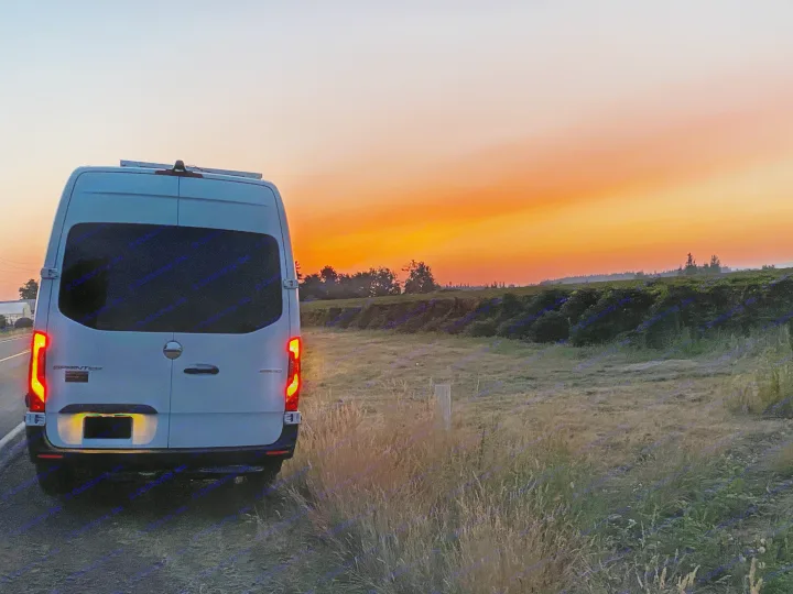 White van, likely an RV rental, parked on a rural road. Its taillights glow against a vibrant orange and red sunset sky.. Freightliner Sprinter 2020