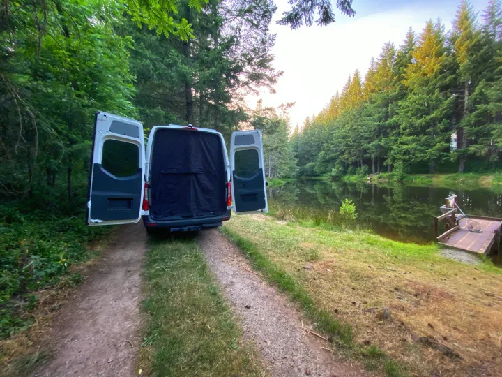 Enjoy nature with this RV rental! Camper van parked by a serene forest lake with open doors, perfect for a peaceful getaway.. Freightliner Sprinter 2020