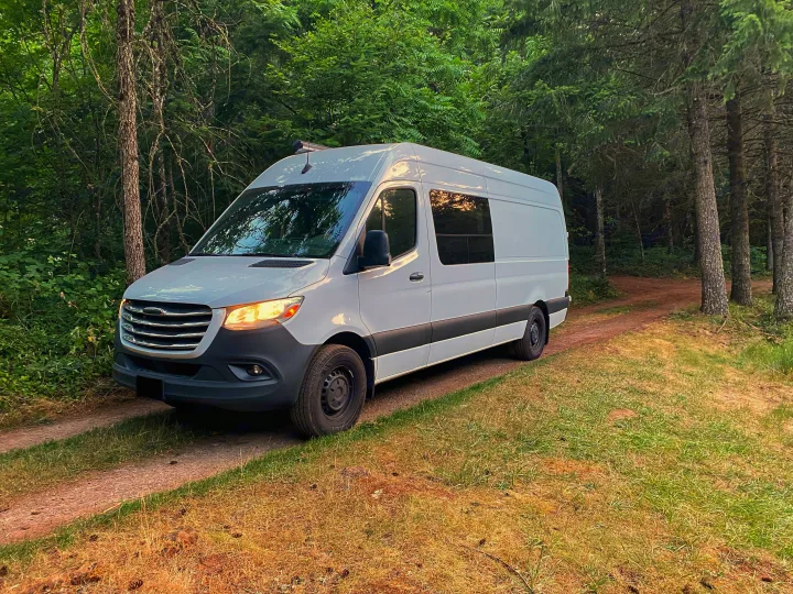 White camper van, an RV rental, parked on a dirt forest path at sunset, perfect for nature getaways.. Freightliner Sprinter 2020