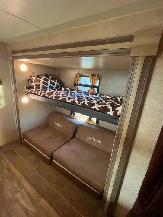 An RV rental's compact bunk area, featuring an upper bed with brown and white bedding, and two lower "Catalina" brown seat cushions.. Coachmen Catalina 2019