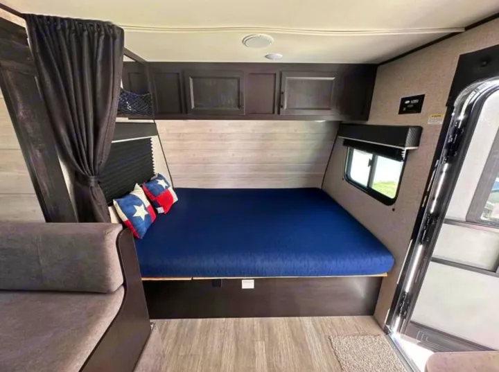 Inside this RV rental: a compact bedroom with a queen bed, blue mattress, Texas flag pillows, overhead cabinets, and a side sofa.. Jayco Jay Flight SLX 2022