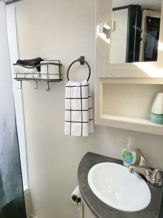 Efficient RV rental bathroom shows compact design: sink, organized shelving for towels/TP, and a forest-themed shower curtain.. Coachmen Apex Nano 2023
